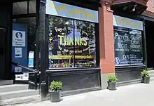 The exterior and sidewalk of a brick building. Above two large windows hangs a sign with the words "THE PUPPET SHOWPLACE." The left window is painted with the words, "Puppet Showplace gives THANKS to our generous donors." The right window is painted with the words, "PUPPETS AT NIGHT. CAMPS. CLASSES. WORKSHOPS." Steps lead up to a glass door through which the Puppet Showplace Theater logo is visible.