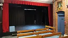 There is a black, proscenium-style stage platform surrounded by bright red curtains. In the foreground are benches with blue cushions. On the right is a brick wall with blue door.