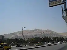 A photo a modern city with a mountain in the background. There is a road, cars, street lights, a sign, and other city infrastructure in the foreground.