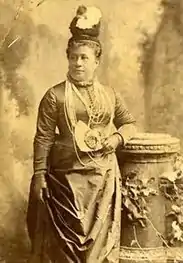 Photograph of Queen Kapiʻolani standing looking to her left. She is wearing  a hat and a necklace of several strands of Ni'ihau shells.