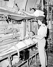 Black and white photo of a woman wearing military uniform standing next two men lying in bunk beds