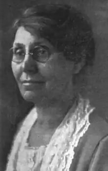 A middle-aged white woman wearing glasses and a lace-collared dress or jacket