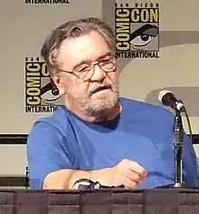 Head and upper torso view of a lightly-bearded, middle-aged man in glasses and a plain T-shirt, sitting behind a table with a microphone on it.  Two small posters that read "Comic Con International" hang on the wall behind.