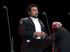 Tenor Ramón Vargas is standing in front of a black backdrop, wearing a tuxedo. Next to him are microphones.