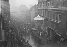 Black and white photo of people wearing military uniforms marching in close formation down a city road. Other people wearing military uniforms are standing on both sides of the road.