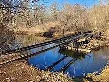 Remains of a railway bridge over a wide stream. Half the bridge structure is gone, leaving just the rails spanning the stream.