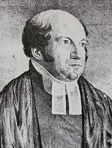 The Reverend Richard Hill is a chubbier man of middle age, with a very bald head fringed with dark curling hair. He wears parson's bands (two white tags at his neck).