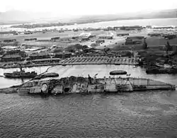 A ship on its side halfway underwater with cables strung to winches on the island behind it.