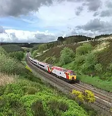 A train in a cutting surrounded by an upland landscape. The tunnel is in the distance behind the train.