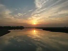 Photograph of the sun very low in the sky, above a wide river with a bridge in the distance. The orange sky and sun are reflected in the river, and the riverbanks are black