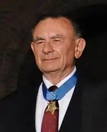 Head of a smiling white man wearing a suit, red tie, and a medal hanging from a blue ribbon around his neck