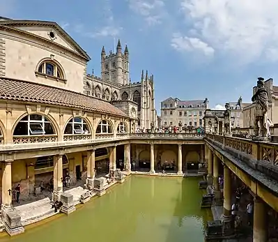 Photograph of the Baths showing a rectangular area of greenish water surrounded by yellow stone buildings with pillars. In the background is the tower of the abbey.