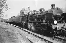  A side-and-front view of a 2-6-0 steam locomotive about to depart a railway station. The locomotive features smoke deflector plates either side of the boiler and there is a crew member oiling the motion. It is hauling four carriages.