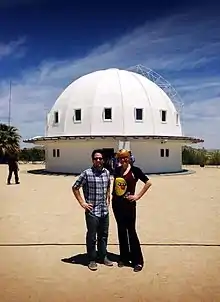 Man with light skin and dark hair and woman with light hair and light skin stand in front of a white building with a domed roof