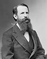 A bearded man sitting down, while wearing old-fashioned formal attire. His prominent beard covers a bow tie.