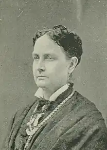 B&W portrait photo of a middle-aged woman with dark hair in an up-do, wearing a white blouse, dark scarf, and dark jacket.