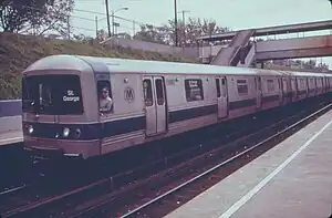 A Staten Island Railway train composed R44 subway cars on the Staten Island Railway. This image, taken in 1973, shows the cars with a since-removed blue stripe toward the bottom of the car body. The train is arriving at a platform to the left; the photo is taken from another platform to the right and in the foreground. The station is in a right-of-way below street level, and a covered footbridge connecting the two platforms is located to the right.