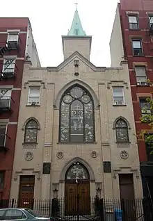 The front of a modest 3-story beige church, between 2 brick apartment buildings, seen from across the street, looking upward. A steeple is visible toward the back.