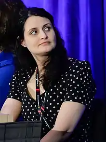 Photograph of a woman sitting at a table on a stage, looking to the side.