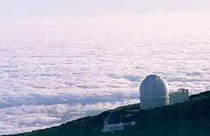 Aerial view of white domed building on side of mountain with floor of white clouds extending to the horizon below and behind the mountain.