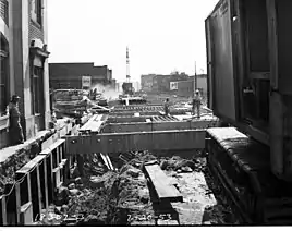 Construction workers observe an open trench formed for a tunnel next to a brick-and-stone building. A concrete and steel framework is being placed over the open trench.