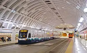 A train in a large, cavernous underground station. A bus can be seen in the background heading the opposite direction.