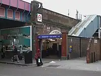 A railway on a brick viaduct crosses a road on a steel bridge, with an entrance below a blue sign reading "SHEPHERD'S BUSH MARKET STATION" in white letters