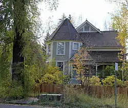 A wooden house with a conical tower in front. There is a tall tree on the left, and many shrubs in front, behind an unpainted wooden picket fence and overgrown grass