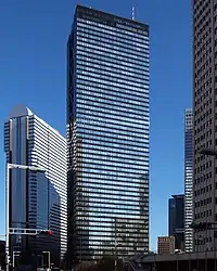 Ground-level view of a black, rectangular high-rise. its glass facades are highly reflective and the smaller facade is bisected by black, inset, crisscrossed beams