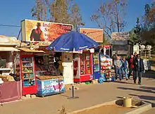 Two small shops offering Coca-Cola products and other soft drinks in glass-fronted refrigerated containers. In the middle is an umbrella with the Pepsi logo. Above them are signs referencing Indiana Jones and depicting him. A small group of people is walking past and in front of them on the right