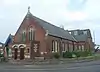 Three-quarter view of a building in red brick and with doubled entrance doors and brick-dressed three-light lancet windows with stone mullions.