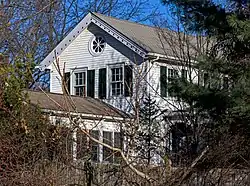 Half of a white wooden clapboard-sided house with a large rose window and decorative vergeboard, seen through an opening in bare-branched shrubbery and evergreens