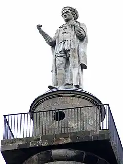 a stone statue of a man in Tudor clothes and down and cap and cahins off office holding a rolled up copy of maga carter