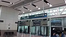 Interior of a modern airport terminal building with glass windows at the top.  A sign hanging from the ceiling reads "skytrain Station 3 / D26-D39, E1-E35".  Passengers are boarding/alighting a train in the bottom right corner.