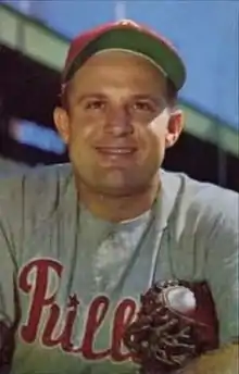 A man wearing a red baseball cap and a gray baseball jersey with "Phillies" written across the chest in red script while clutching his baseball mitt under one arm