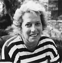 A black and white portrait photo of a smiling woman with curly hair. She is seated and wearing a striped top.