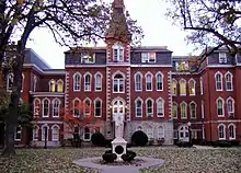 A statue sits in front of a large four-story red brick building. To the right of the entrance, the building goes in a little ways, and then back out. To the left of the entrance is a small two-story area connecting the main part of the building to the rest of it. Above and behind the two-story part is a four-story part of the building