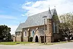 Three bay nave and chancel. No side aisles. Side entry porch and small belfry. Slate roof. Walls dressed stone, brick dressings. Wooden windows. School house/parish hall of similar materials. Built 1861. Type of site: Church. Current use: Religious – Church.