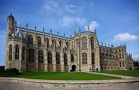 A photograph of a huge stone chapel, with tall stained glass windows and stone detailing. A patch of green grass can be seen in the foreground with several people walking by the door of the chapel.