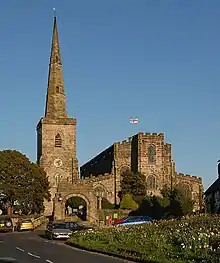 A Gothic stone church with a tall steeple on the left linked to the battlemented body of the church on the right