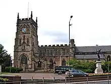 alt=A large medieval sandstone church, with tower, a clock face and stained glass windows.
A statue of an angel of peace in the foreground