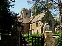 A view through a gate and graveyard to a yellow stone building with a square tower.