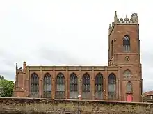 A church seen from the south with a west tower surmounted by a pierced parapet and pinnacles