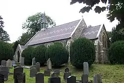 A small church with a decorated roof seen from the southeast. On the west gable is a simple bell tower. In front of the church is a row of small yews and in the foreground are gravestones