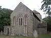 A flint rubble church with large stone quoins.  In the nearest wall is a two-light lancet window between buttresses.  A squat tower with a squared-off pyramidal roof is partly obscured behind this.