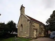 A small simple, stone church seen from the south-west, showing a bellcote, buttresses, and a round-arched doorway and west window, each flanked by columns