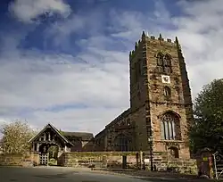 A sandstone church seen from the west with the tower prominent. In front of this is a wall leading to a lych gate on the left. In front of the wall are stocks