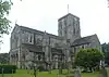 Three-quarter view of a substantial stone church with a square clock tower topped with a weather-vane. The tower has two- and three-light arched openings in each face. A triangular-roofed projection to the right has several round-arched windows. The main body of the church extends to the left and has large buttresses topped with spirelets.