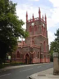 A brick tower with red decorations, including a pierced parapet and crocketed pinnacles