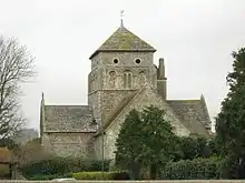 A cruciform church of flint and stone with a pyramid-roofed central tower topped with a weather-vane. The nearest section is fully obscured by a tree. The tower has three blocked round-arched windows and two round openings below the roof.